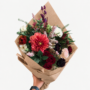 A hand holding a vibrant mixed floral bouquet wrapped in brown kraft paper against a clean white background. The bouquet features a prominent coral-pink dahlia, deep red and burgundy roses, white and pink blossoms, and green foliage.