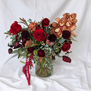 A floral arrangement in a clear glass vase featuring deep red roses, burgundy berries, peach orchids, and green foliage, set against a draped white fabric background.