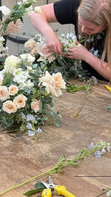 Several women arranging floral arrangements with white and cream roses, green foliage, and light blue flowers at a rustic wooden table.