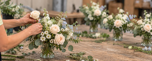 A florist arranges a bouquet of pale pink roses, white daisies, and light blue filler flowers with green foliage in a glass vase on a rustic wooden table. Other bouquets and floral tools are visible in the softly blurred background.