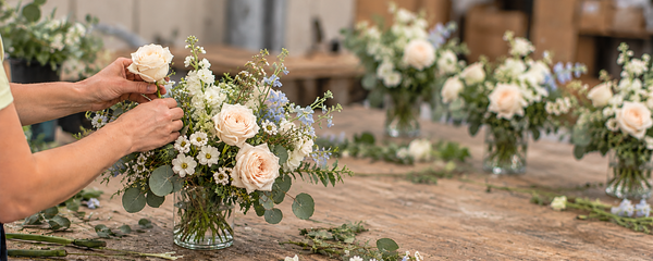 A florist arranges a bouquet of pale pink roses, white daisies, and light blue filler flowers with green foliage in a glass vase on a rustic wooden table. Other bouquets and floral tools are visible in the softly blurred background.