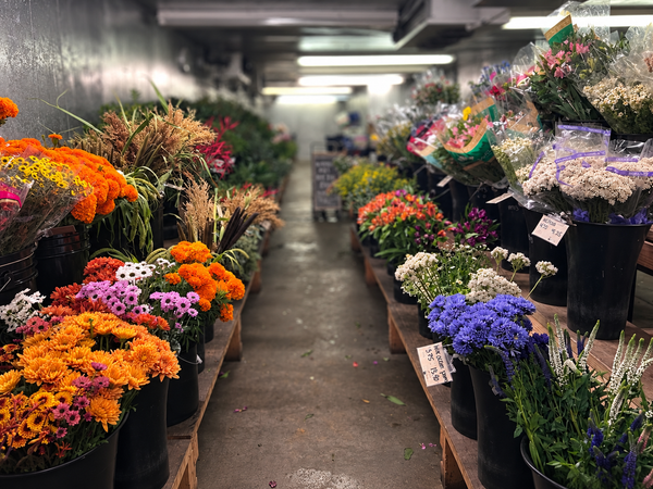 A narrow aisle in a flower market  lined on both sides with black buckets filled with colorful flowers, including orange, purple, white, and yellow blooms.
