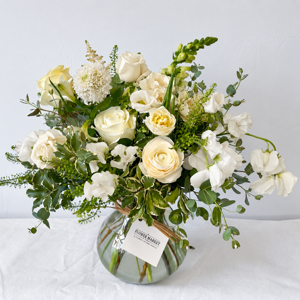 A floral arrangement of white and cream roses, hydrangeas, and green foliage in a clear glass vase with a striped base, set against a plain light background. A small tag on the vase reads Flower Market.