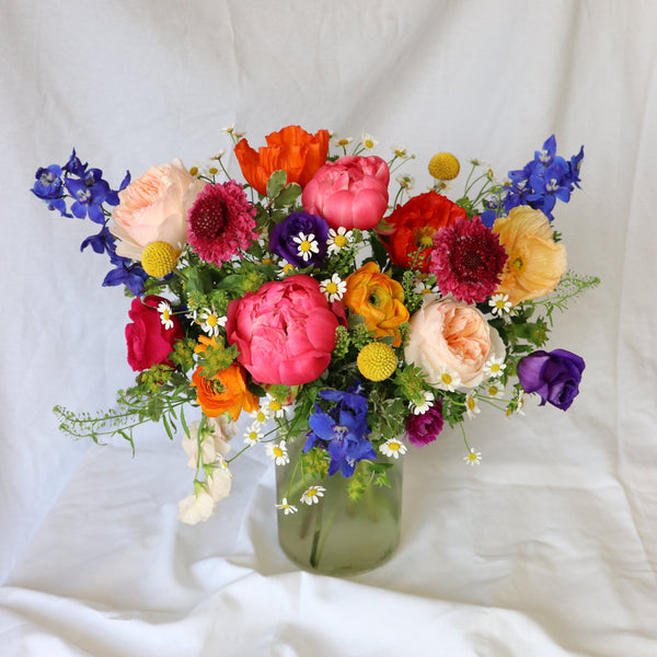 A vibrant floral arrangement featuring a variety of bold blooms including peonies, garden roses, ranunculus, tulips, delphinium, scabiosa, lisianthus, and sweet pea in a ceramic compote vase.