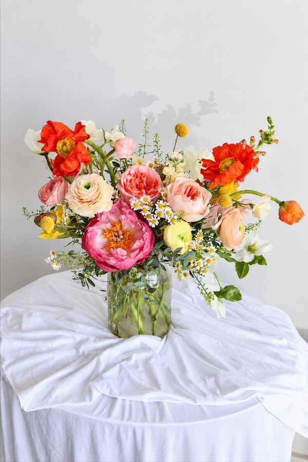 A vibrant, multicolor floral arrangement featuring pink, red, peach, and white flowers with green foliage, presented in a clear glass jar on a white draped table against a light gray wall.